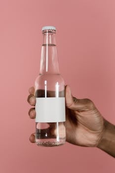 Close-up of a hand holding a glass bottle with a blank label on a pink background.