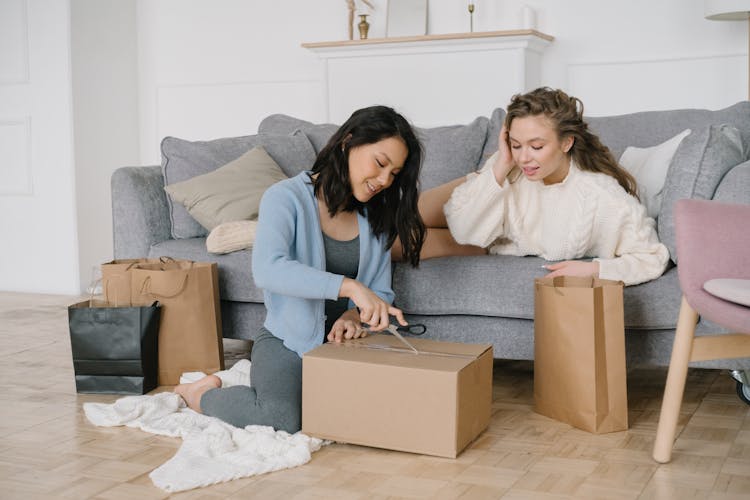 A Woman In Knitted Sweater Looking At The Other Woman Opening The Cardboard Box Using Scissors