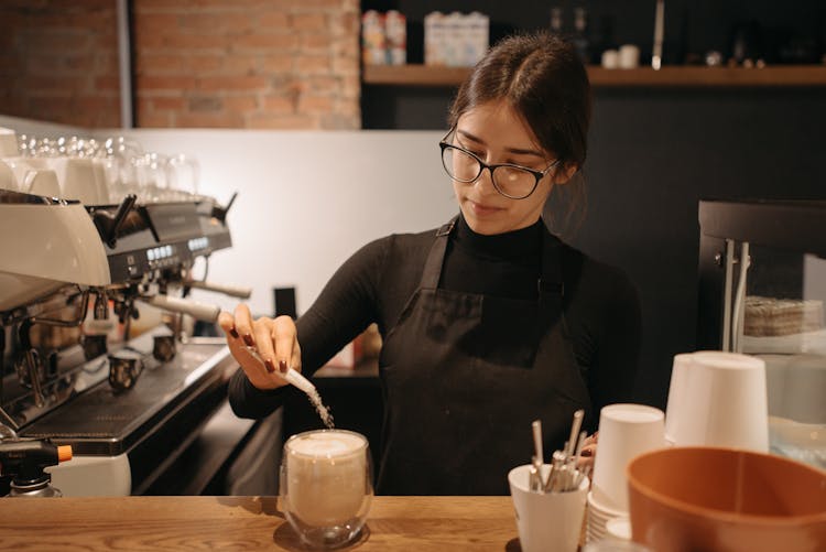 A Woman Pouring A Sachet Of Sugar In A Glass Of Coffee At The Counter