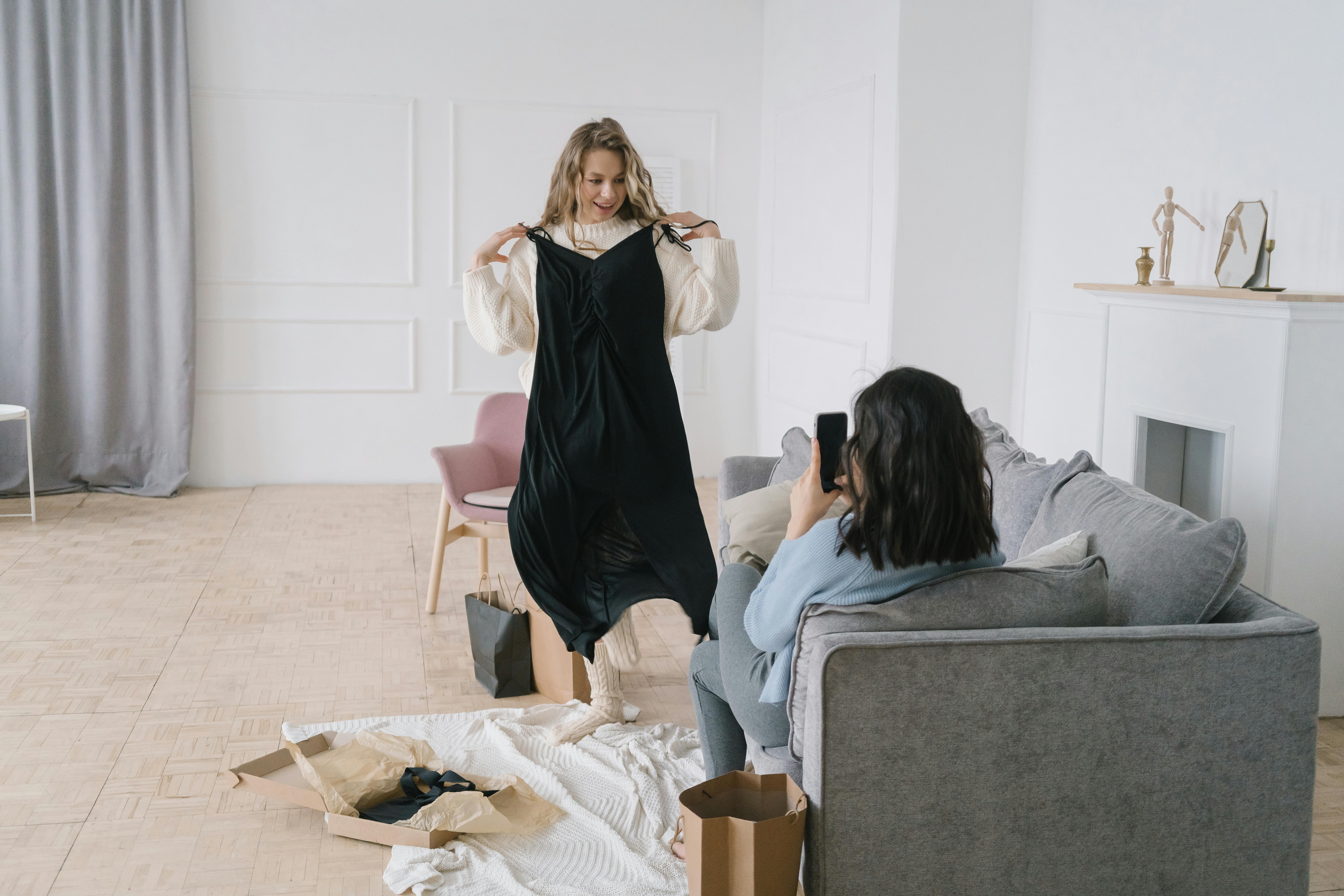 Young woman tries on a black dress in a cozy living room, captured by a friend.