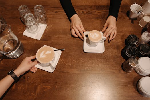Two baristas serve lattes with foam art on a wooden table in a cozy coffee shop.