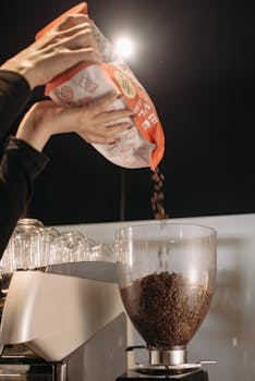 A barista pours coffee beans into a grinder, preparing for brewing in a stylish coffee shop setting.