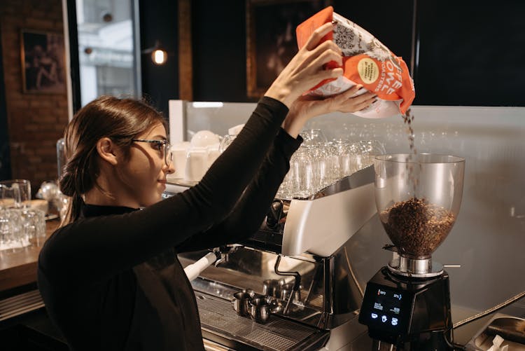 A Woman In Black Long Sleeves Pouring Coffee Beans In A Grinder