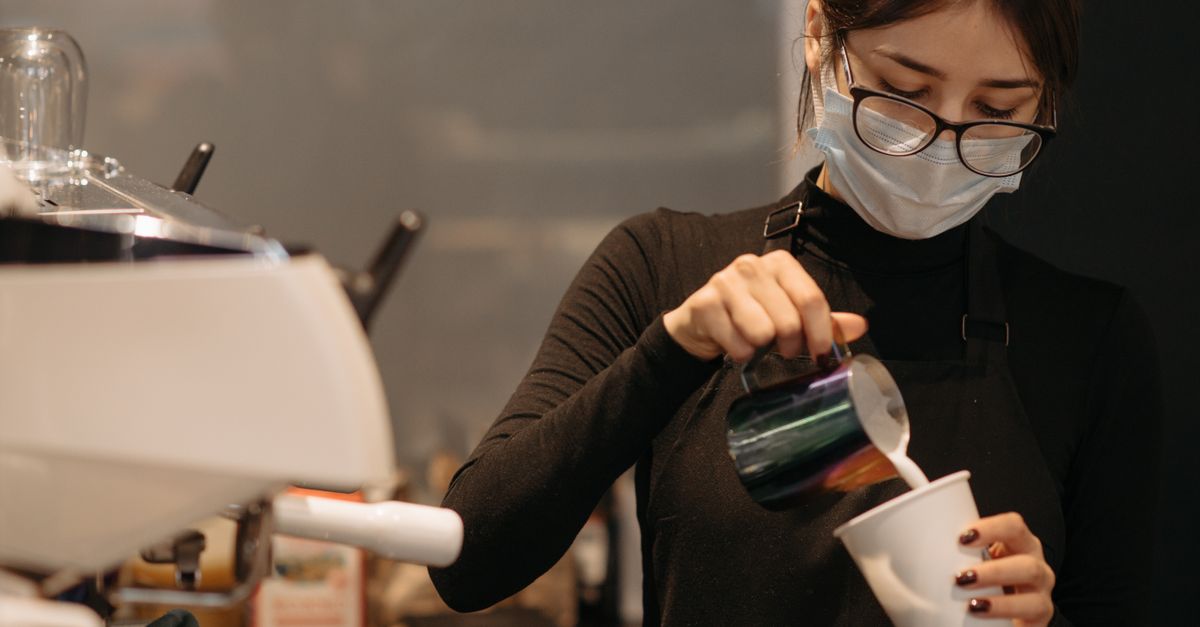 A focused barista wearing a mask pours milk into a coffee cup at a café.