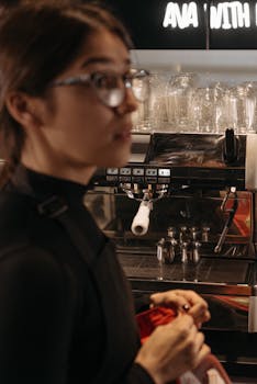 A barista with eyeglasses stands beside an espresso machine in a cozy cafe setting.