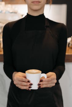 A barista in a black apron and turtleneck holds a steaming cup of coffee indoors.