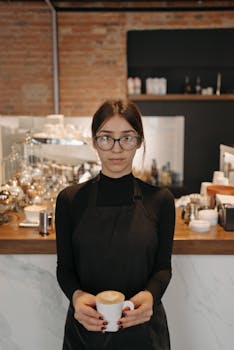 A barista in a black apron and eyeglasses holds a cup of coffee in a warm cafe environment.