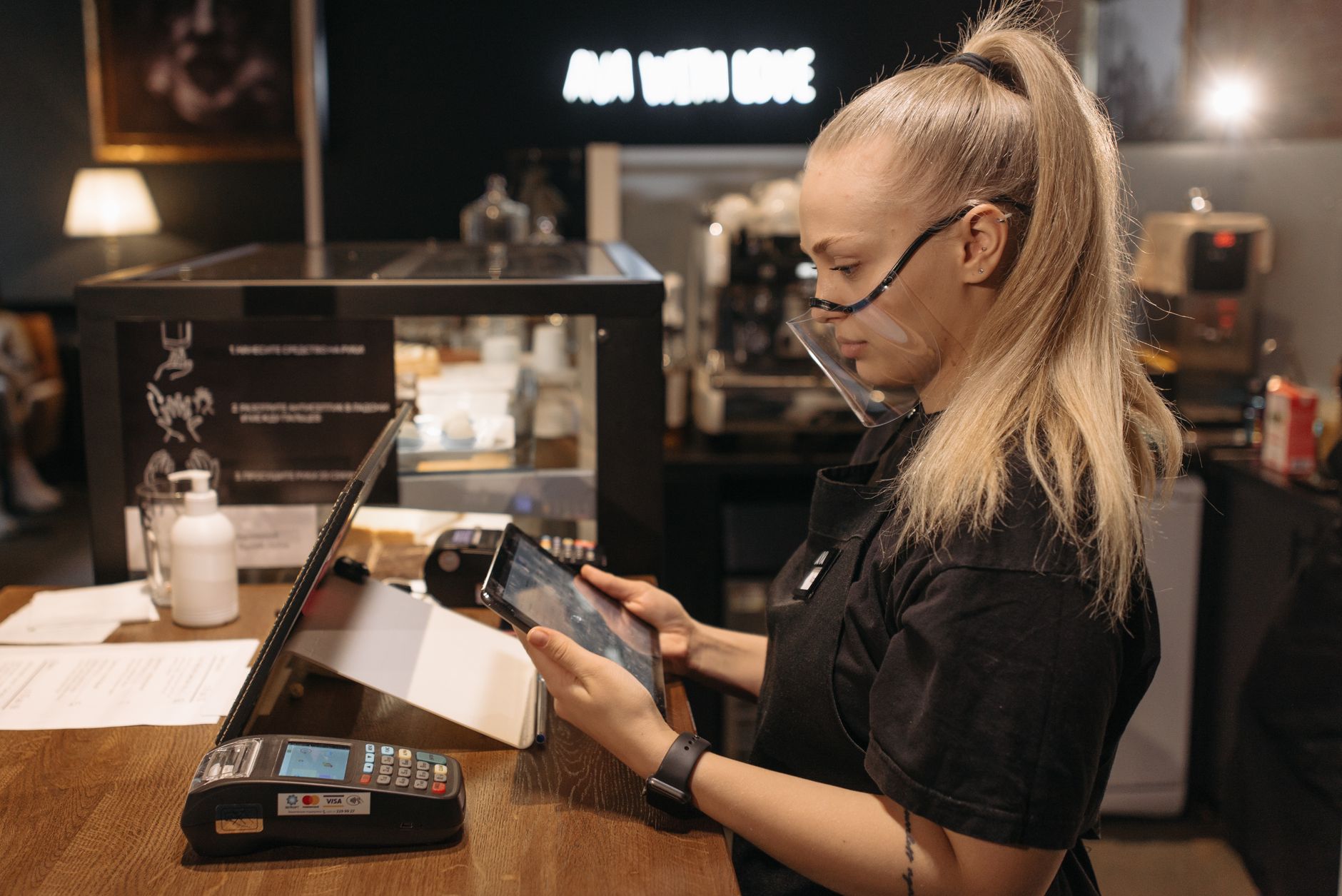A barista wearing a face shield uses a tablet for work at a coffee shop counter, emphasizing modern service.