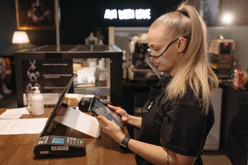 Free A barista wearing a face shield uses a tablet for work at a coffee shop counter, emphasizing modern service. Stock Photo