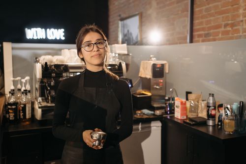Free Smiling barista holding portafilter inside a cozy cafe with coffee machine, wearing apron and glasses. Stock Photo