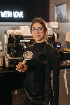 Barista with eyeglasses and apron smiling while holding a portafilter in a cozy coffee shop.