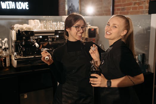 Two baristas smiling and standing together in a café holding coffee tools.