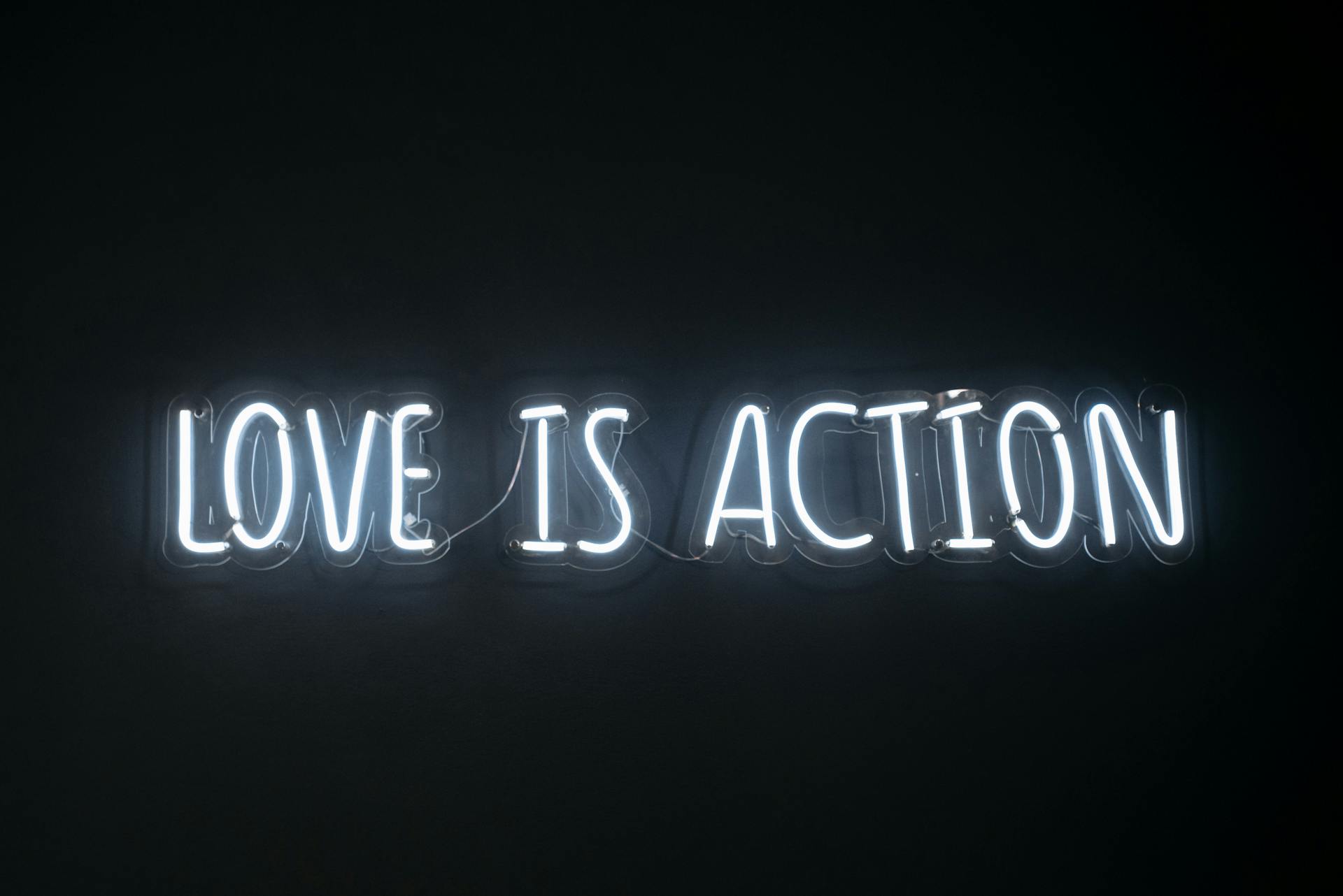 Bright neon sign reading 'Love is Action' against a dark background.