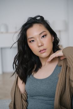 A young woman with dark hair poses confidently indoors wearing a tank top and cardigan.