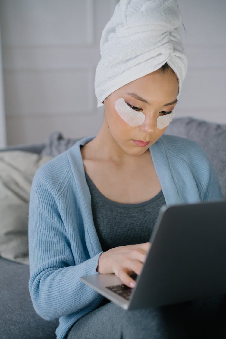 Woman In Blue Sweater With Head Towel And Under Eye Mask Using Laptop