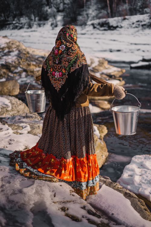 Free A woman in colorful attire carrying metal buckets near a snowy river. Stock Photo