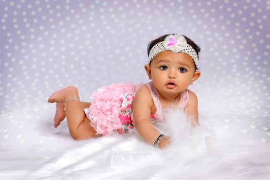 Full body of adorable little ethnic girl wearing headband and colorful dress looking away while lying on soft white textile in light room