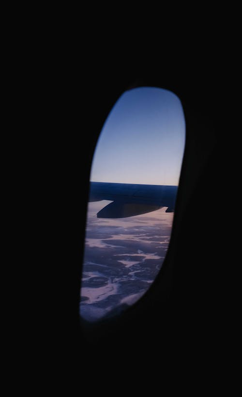 Through window of wing of modern aircraft flying in blue sky over mountains and lakes at sunset