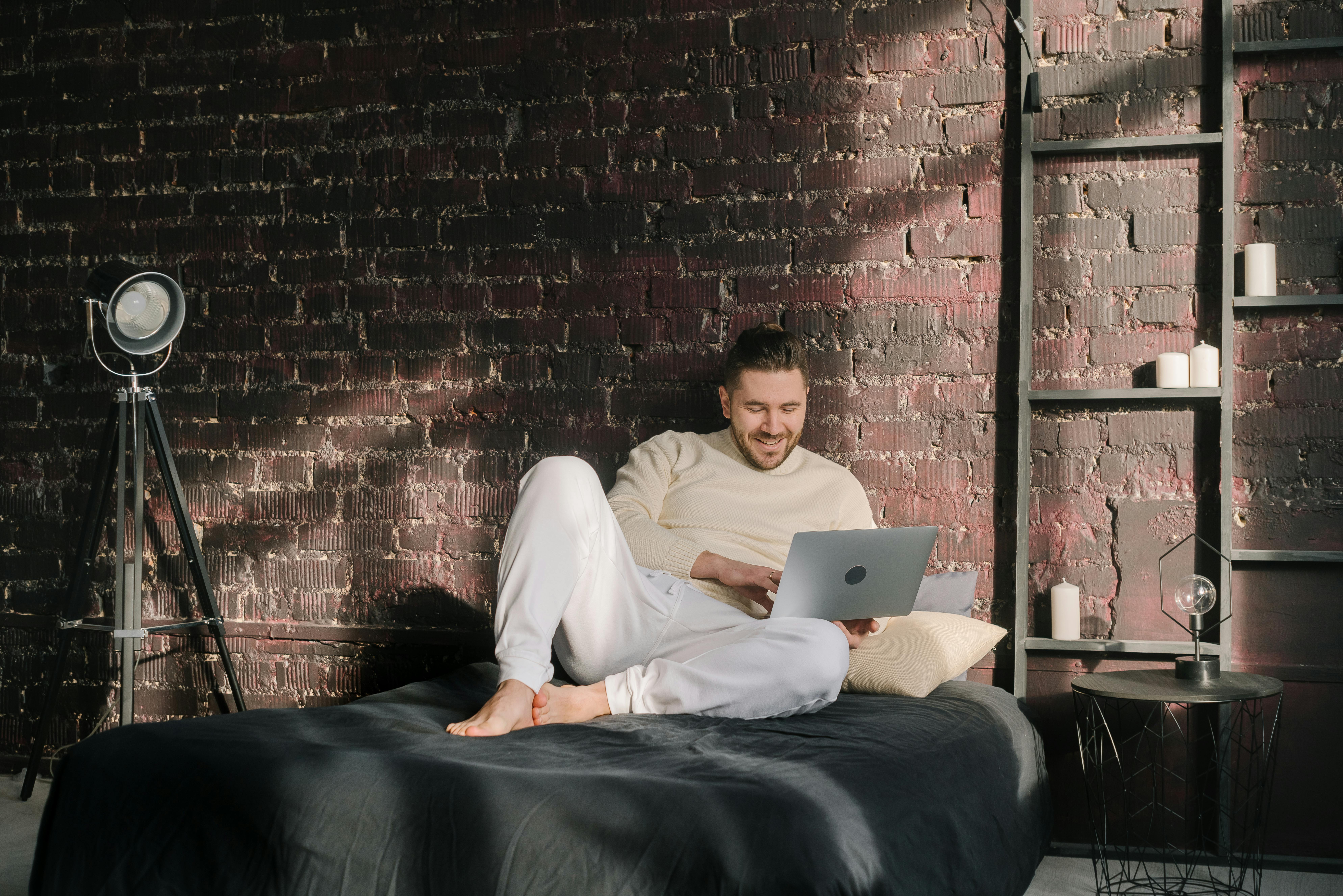Man reclining on a bed using a laptop in a cozy room, smiling while making a video call.