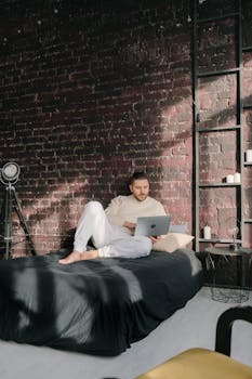 Man comfortably working from bed with a laptop in a stylish, sunlit room.