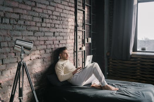 Young man enjoying remote work on laptop in a modern industrial bedroom setting.