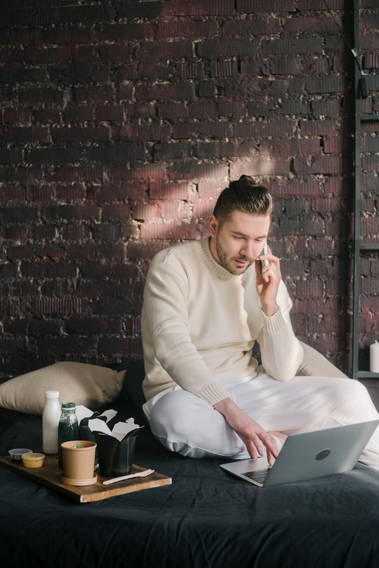 Man In White Sweater Sitting On Bed Using Laptop And Cellphone Beside A Take Out Food