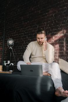 Man in casual attire engaged in a remote work setup, seated on a bed with a laptop and smartphone.