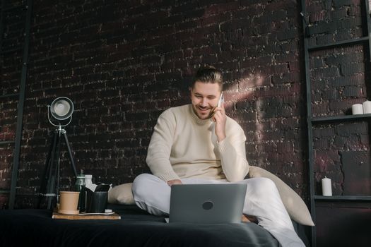A smiling man sitting with a laptop and cellphone in a cozy indoor setting.