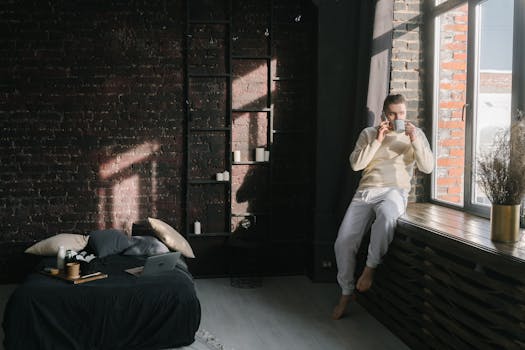 Man enjoying a coffee break by a sunlit window in a cozy bedroom setting.