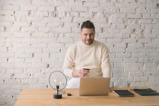 Adult man using credit card and laptop for online shopping in bright, minimalist office setting.