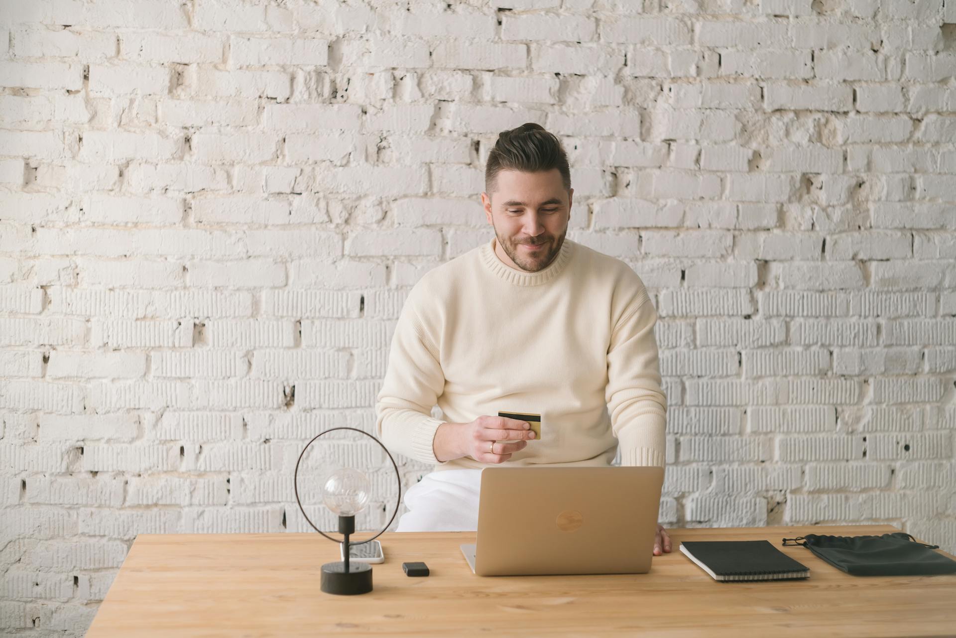 Adult man using credit card and laptop for online shopping in bright, minimalist office setting.