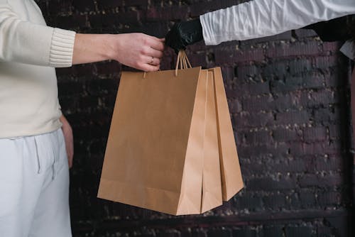 A Person Wearing Black Gloves Handing Paper Bags to Another Person Wearing Beige Sweater