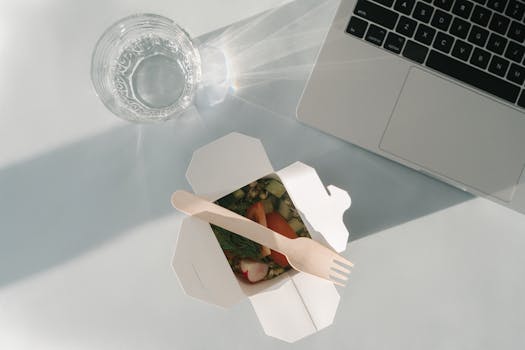 Healthy salad in a takeout box with a glass of water and a laptop on a white surface, top view.