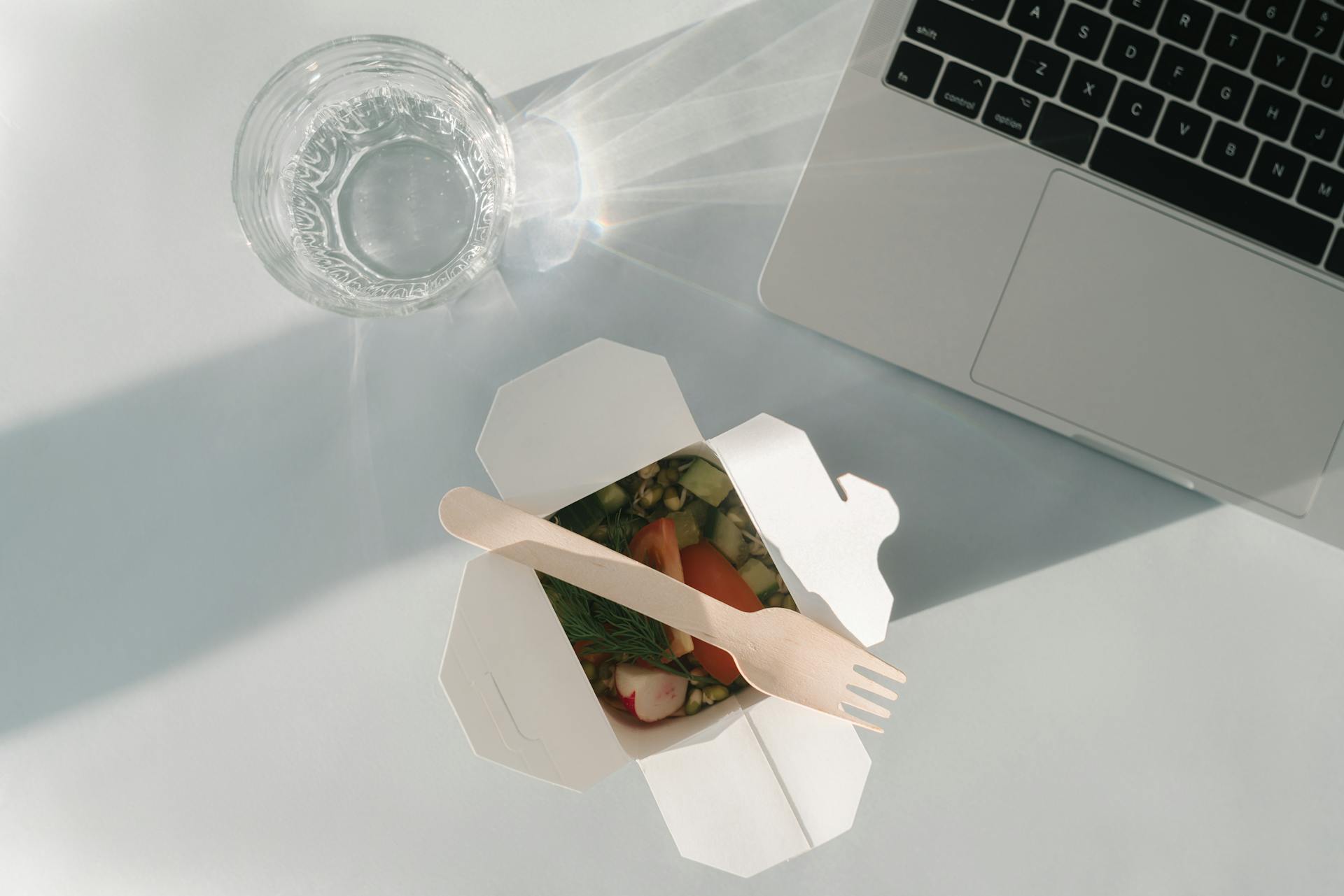 Healthy salad in a takeout box with a glass of water and a laptop on a white surface, top view.