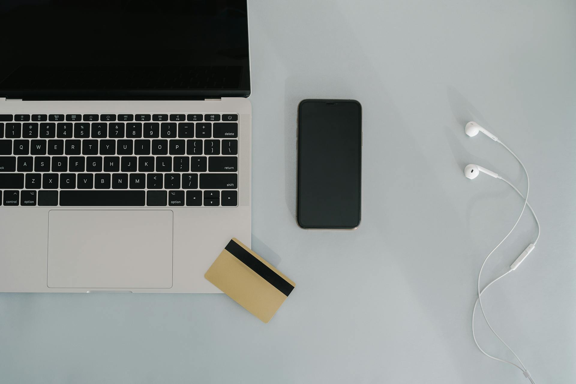 Flat lay of laptop, smartphone, and earphones with credit card on a desk.