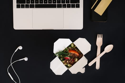 A flat lay of a workspace setup with a laptop, smartphone, earbuds, and a takeout meal.