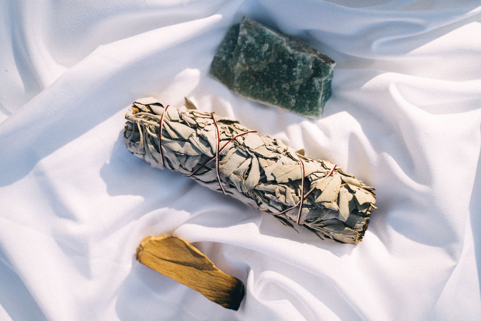 A flat lay of sage, palo santo, and crystal on white fabric, symbolizing cleansing rituals.