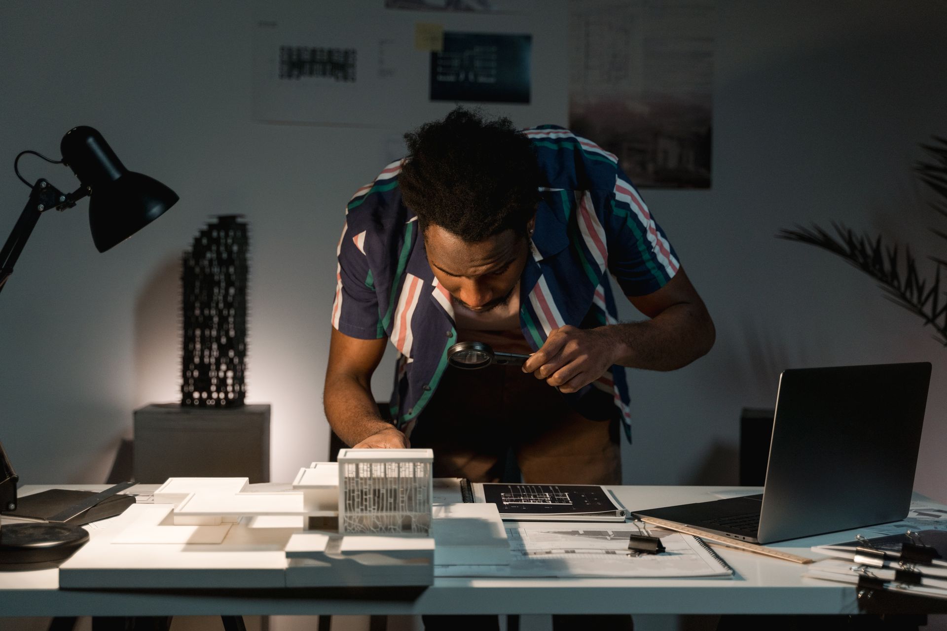Focused architect analyzing detailed model with magnifier, working late indoors.