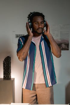 A man with eyes closed, wearing headphones, listens to music in a cozy indoor setting.