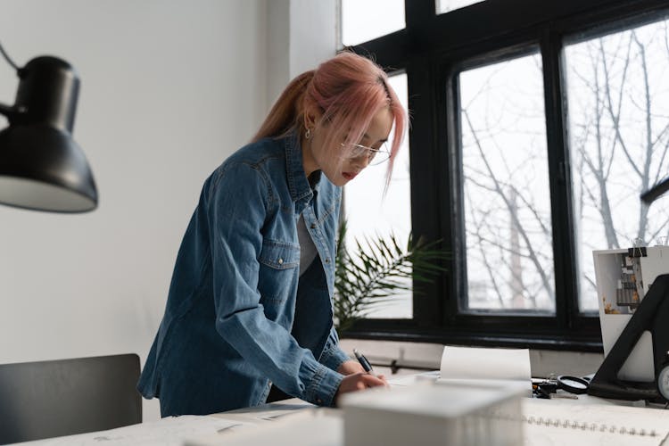 A Woman With Pink Hair Busy Working On Her Table Near Glass Window