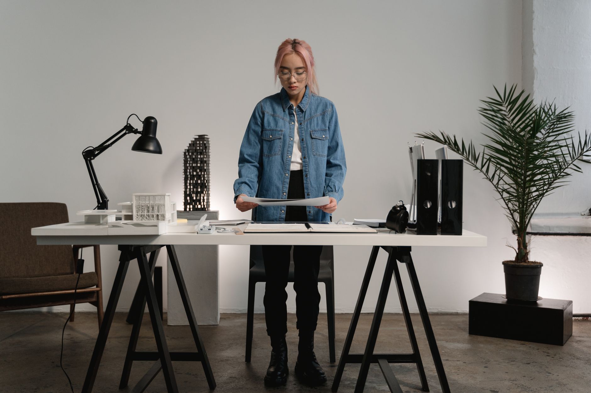 A woman with pink hair stands at a desk in a contemporary office space, examining documents.