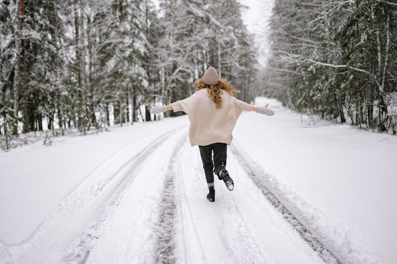 Woman in a cream sweater, knit toque and mittens walking down a snowy forest road