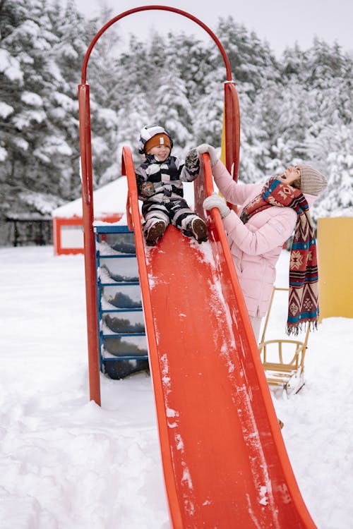A Smiling Child in Snow Suit Sitting on a Red Slide Beside a Laughing Woman
