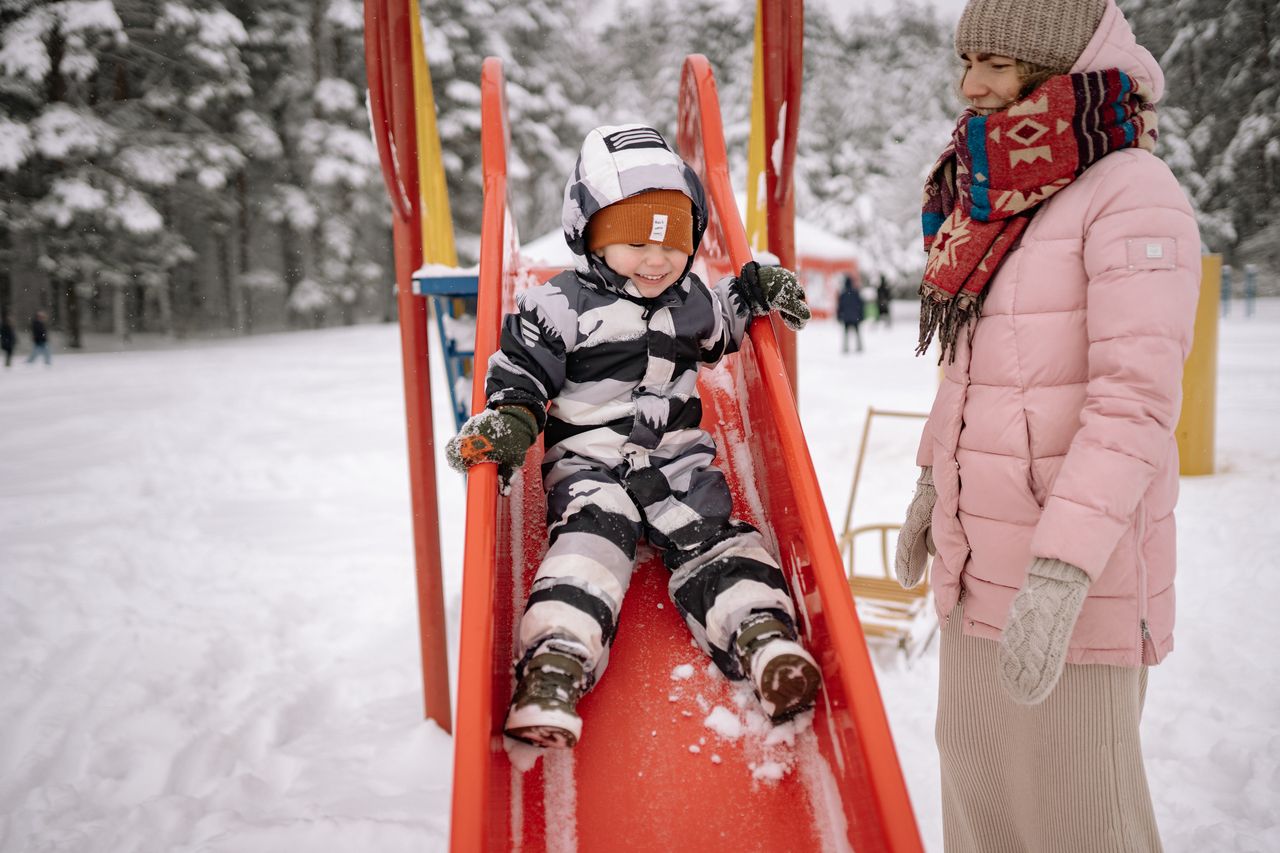 Toddler in a full snowsuit sliding down a snowy playground slide