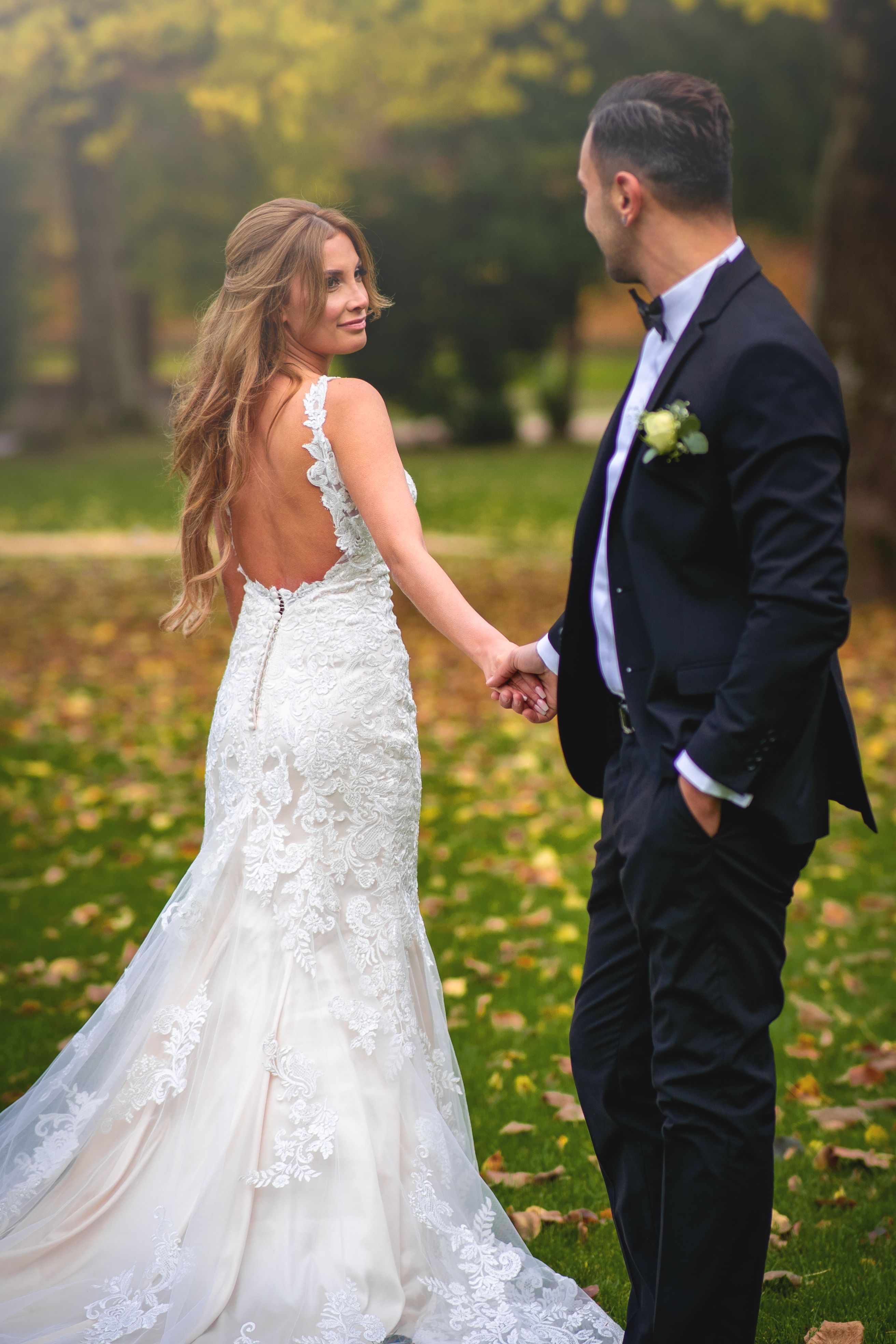 Bride in lace gown holding hands with groom in tuxedo, surrounded by autumn leaves.