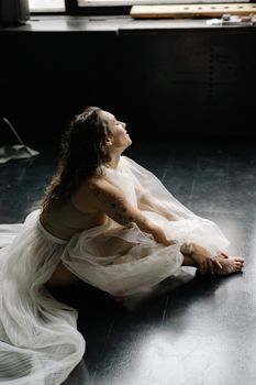 A serene ballerina sitting on a dark wooden floor in a studio setting, enveloped in a light, flowing dress.