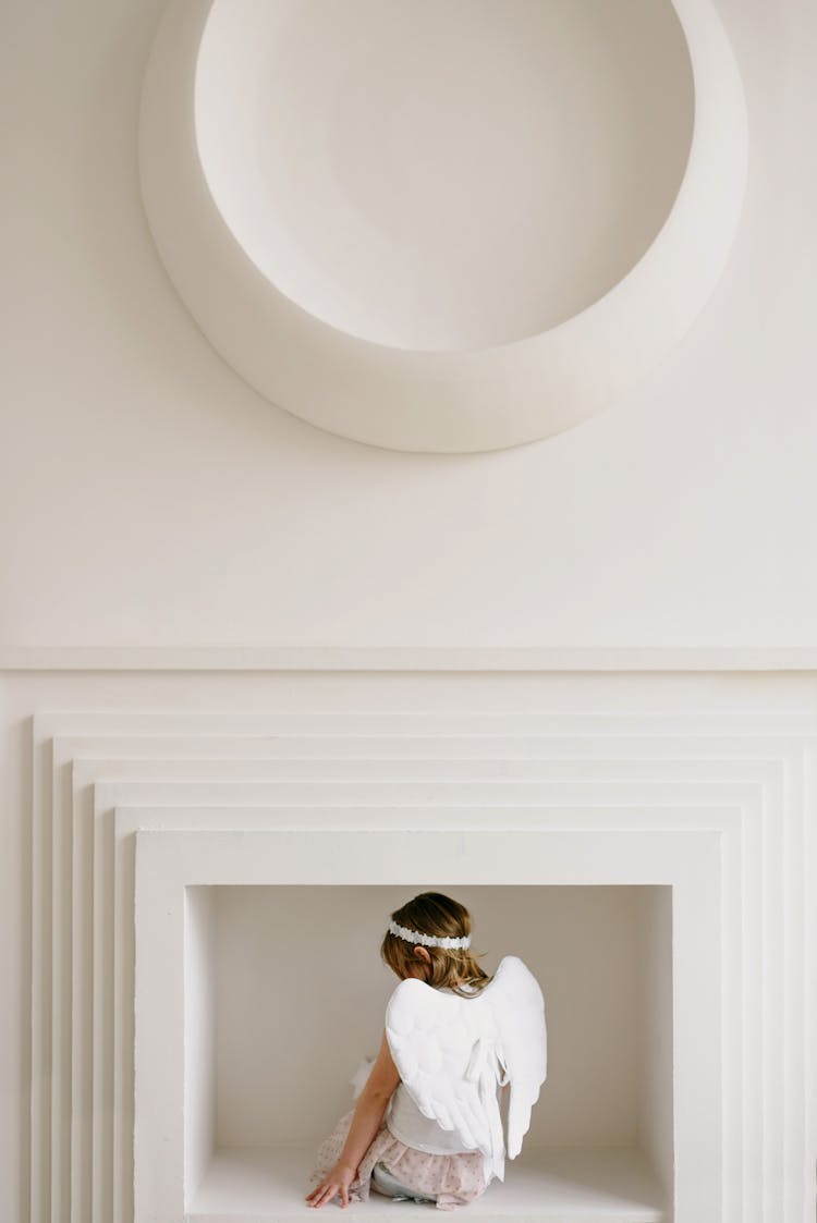 A Girl In An Angle's Costume And Halo Sitting Near A White Wall