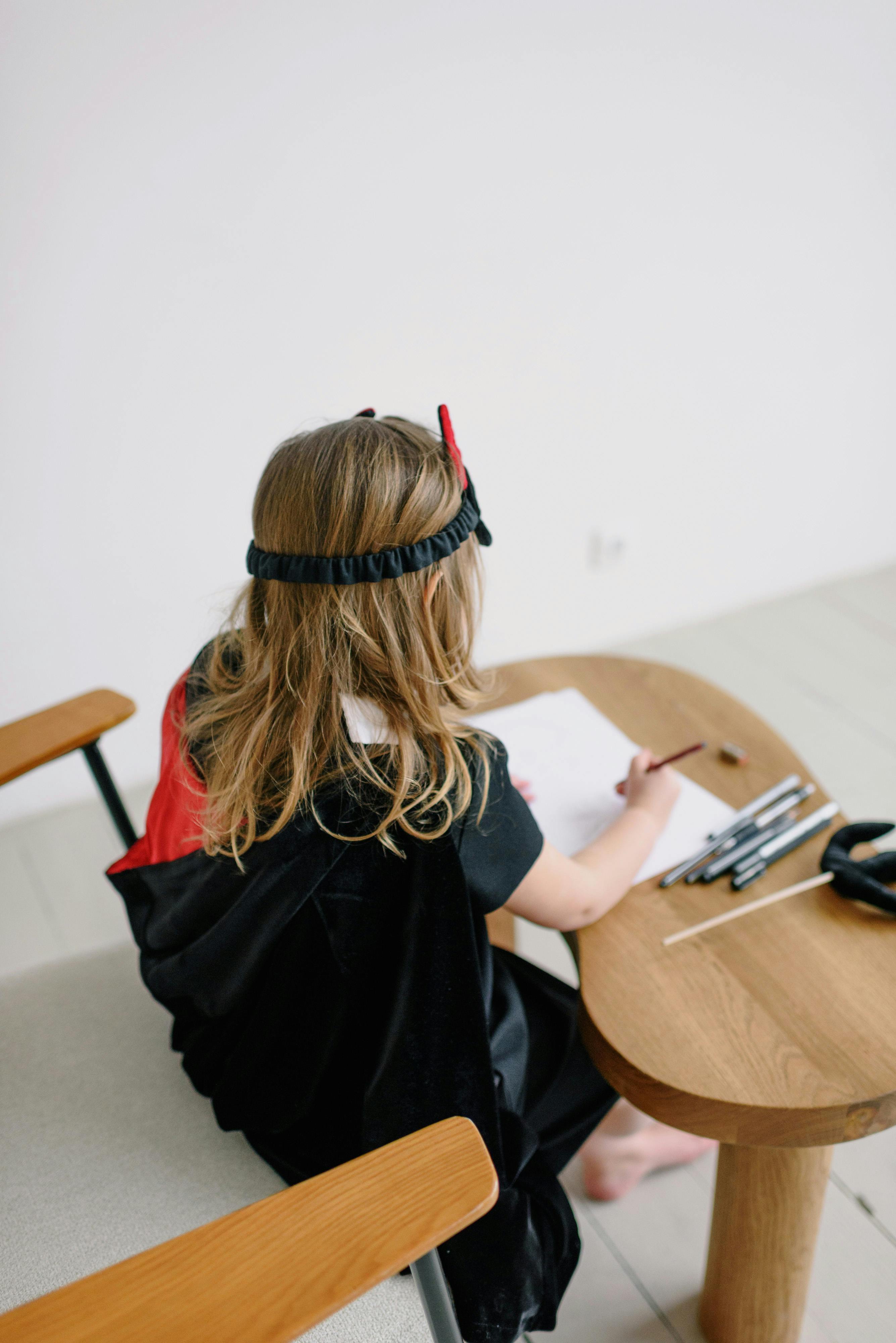 Free A young girl in a demon costume sits drawing on a wooden table indoors. Stock Photo