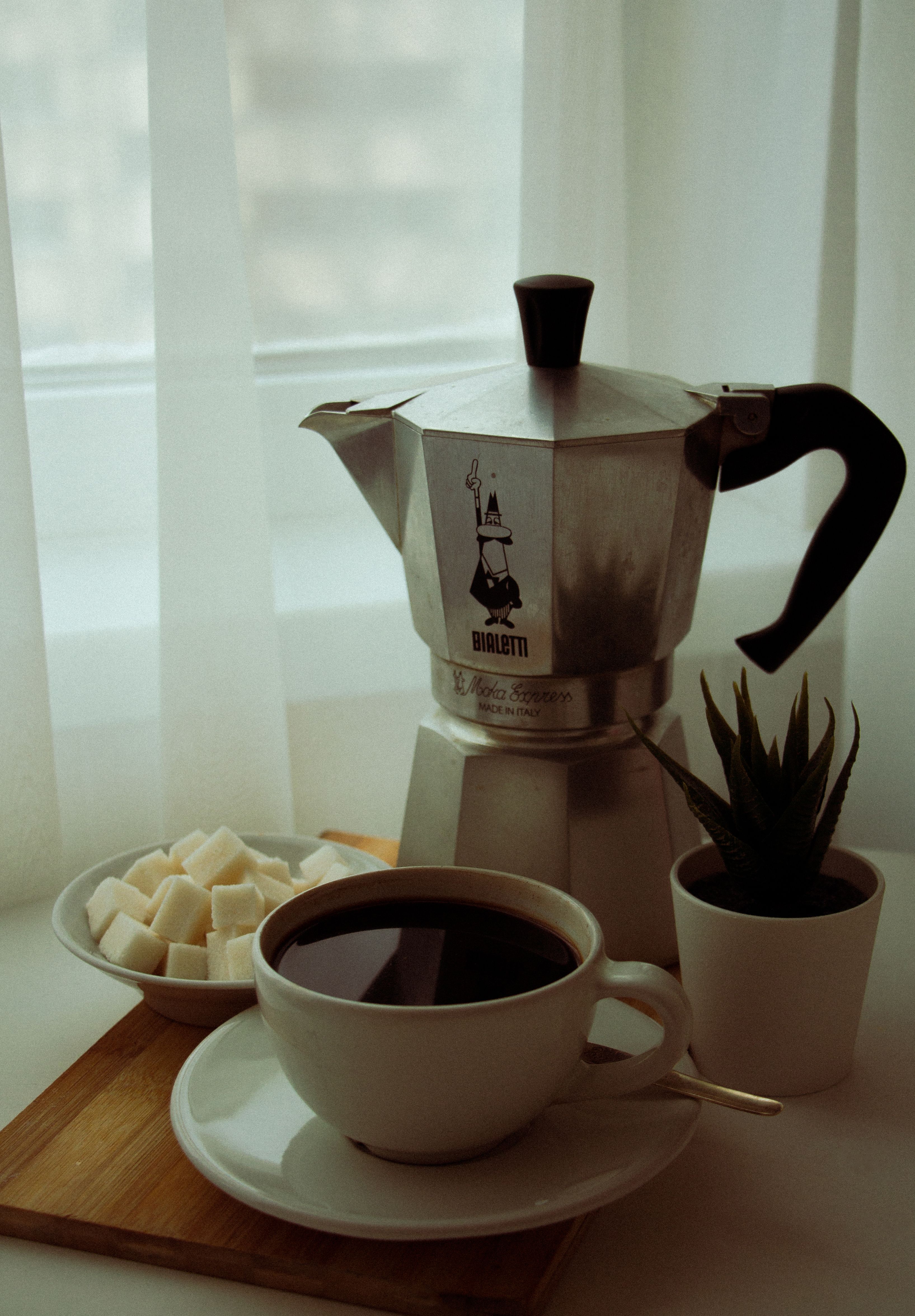 A peaceful morning coffee scene with a moka pot, cup of coffee, and sugar cubes by the window.