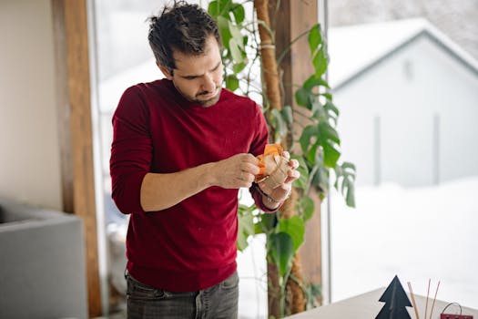 A man in a red sweater wraps a gift in a cozy indoor setting, surrounded by plants. Winter vibes.